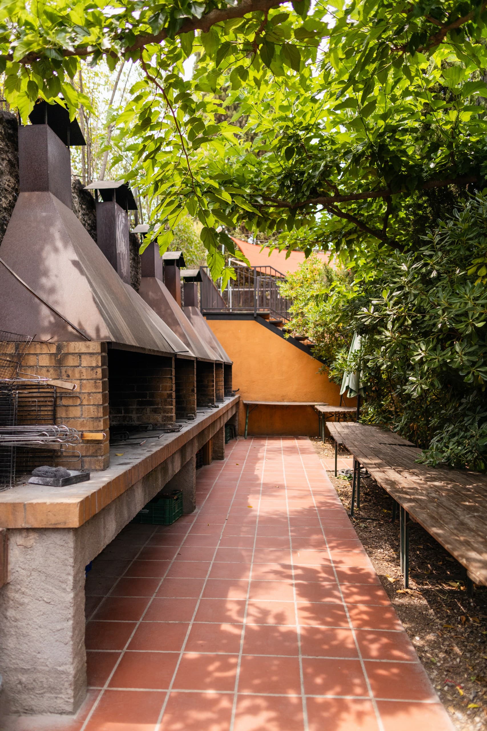 Brick outdoor grills with metal chimneys on a red tiled patio under green trees.