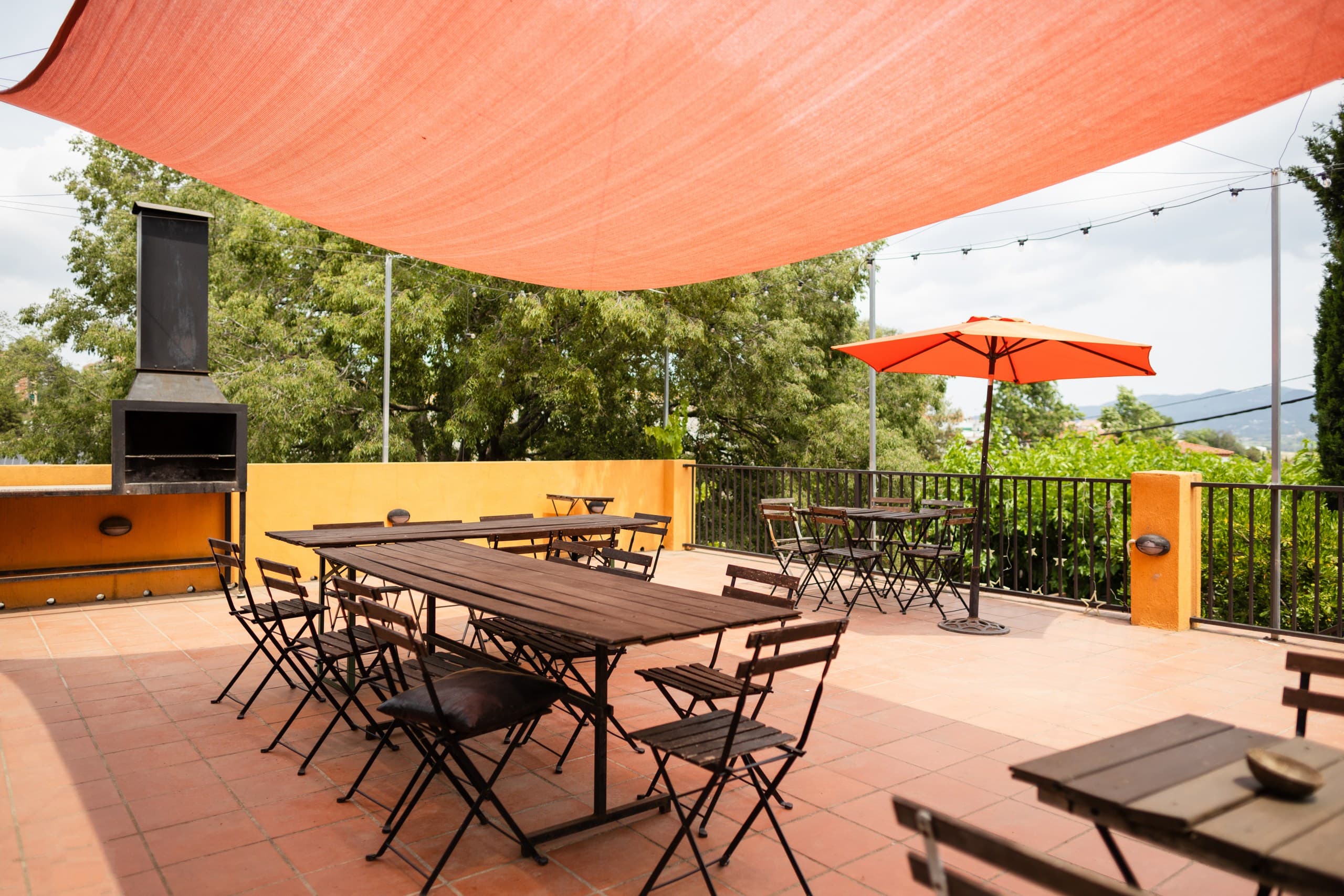 Outdoor terrace with wooden tables and chairs under a large orange sunshade and umbrella.