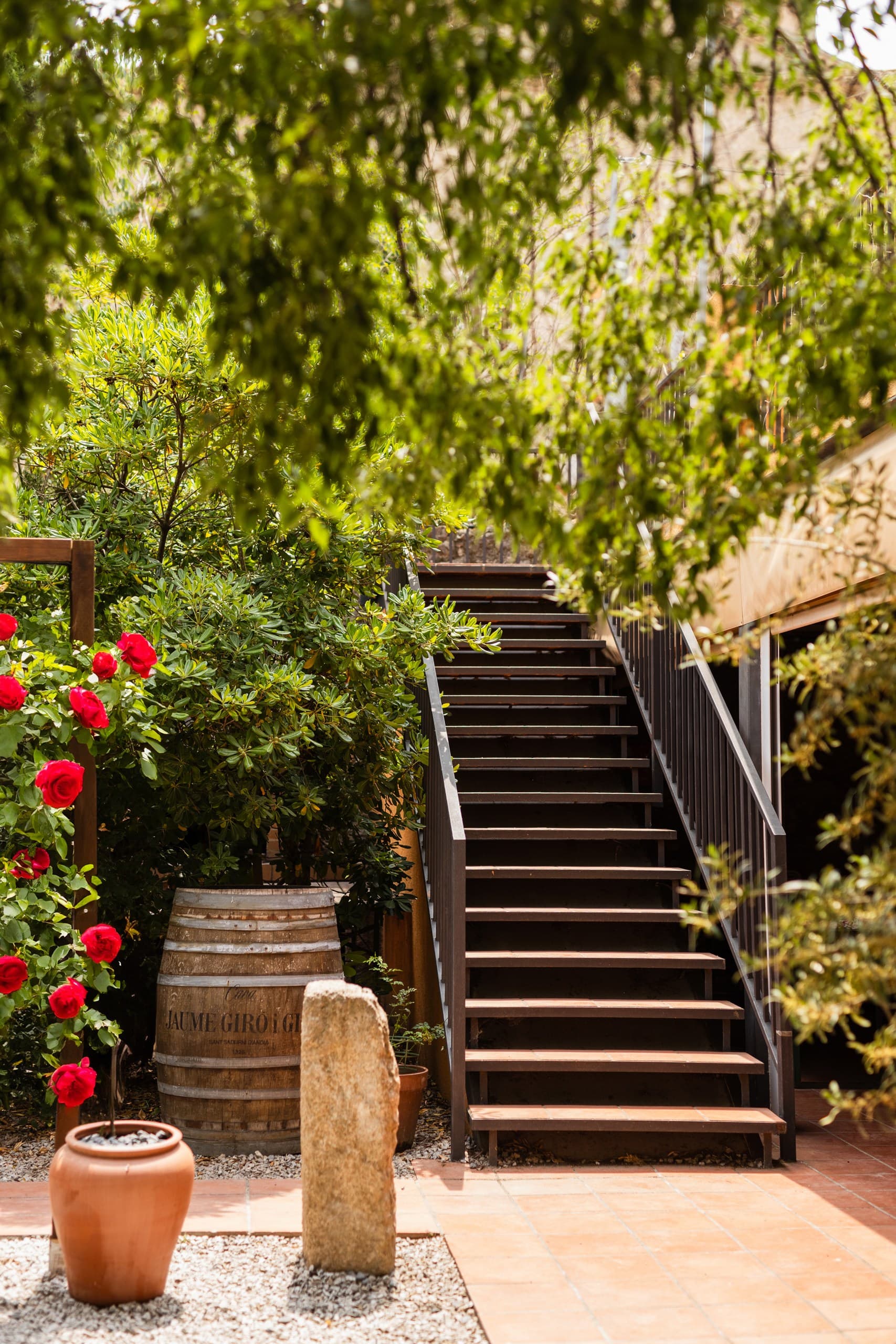 Outdoor wooden staircase ascending through lush greenery, red roses, and a rustic wine barrel.