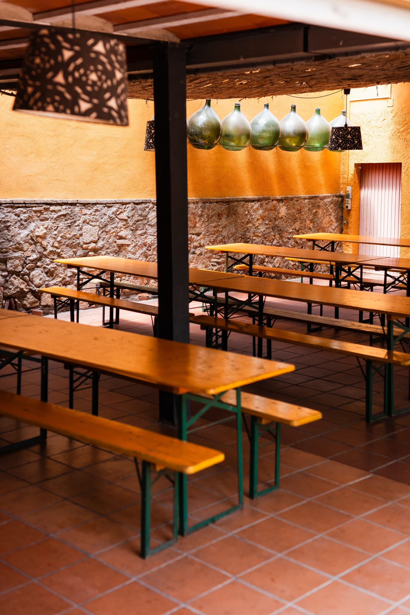 Rustic patio with wooden picnic tables, stone walls, and decorative hanging green glass bottles.