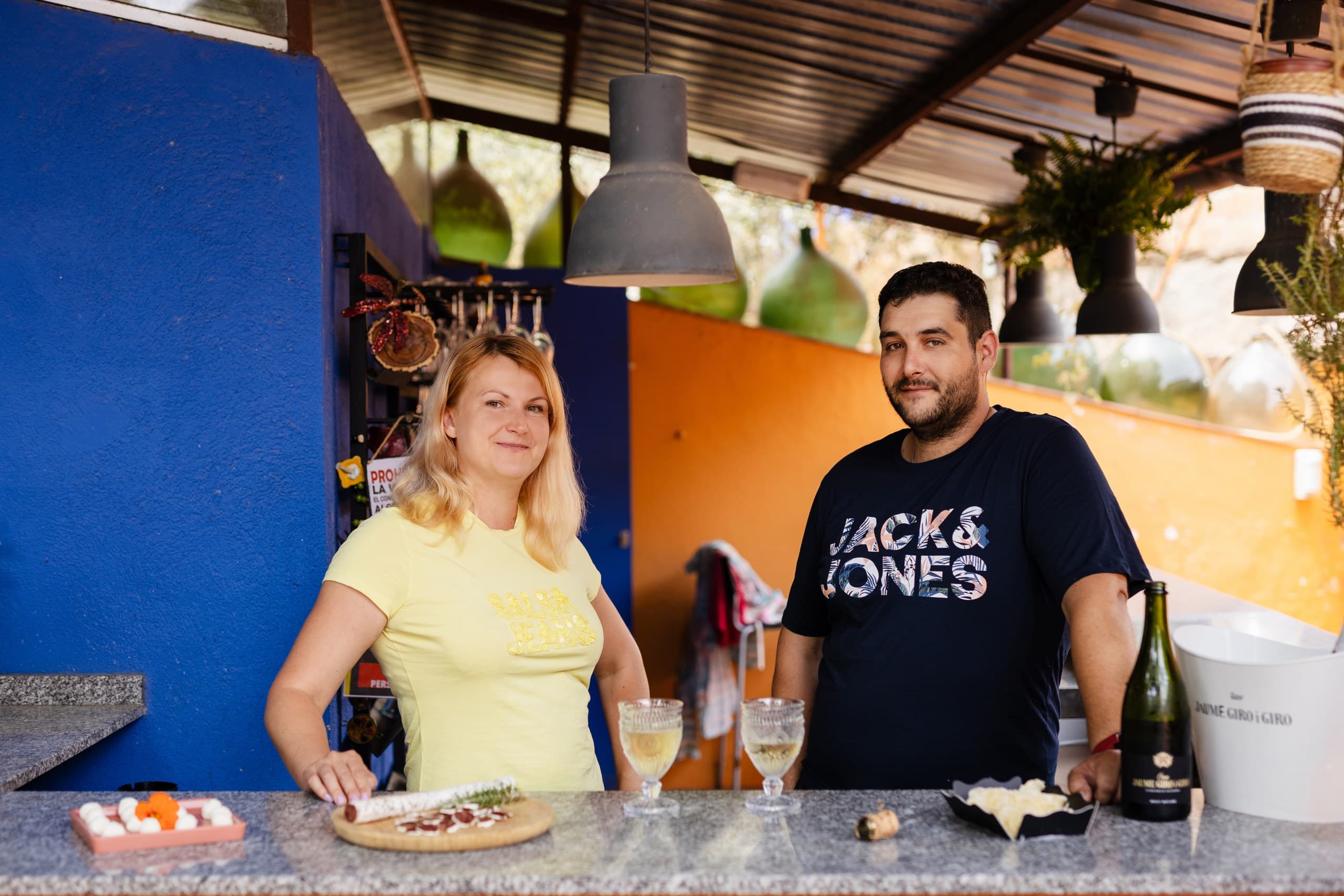 Smiling man and woman behind a bar counter with wine, cheese, and cured meats.