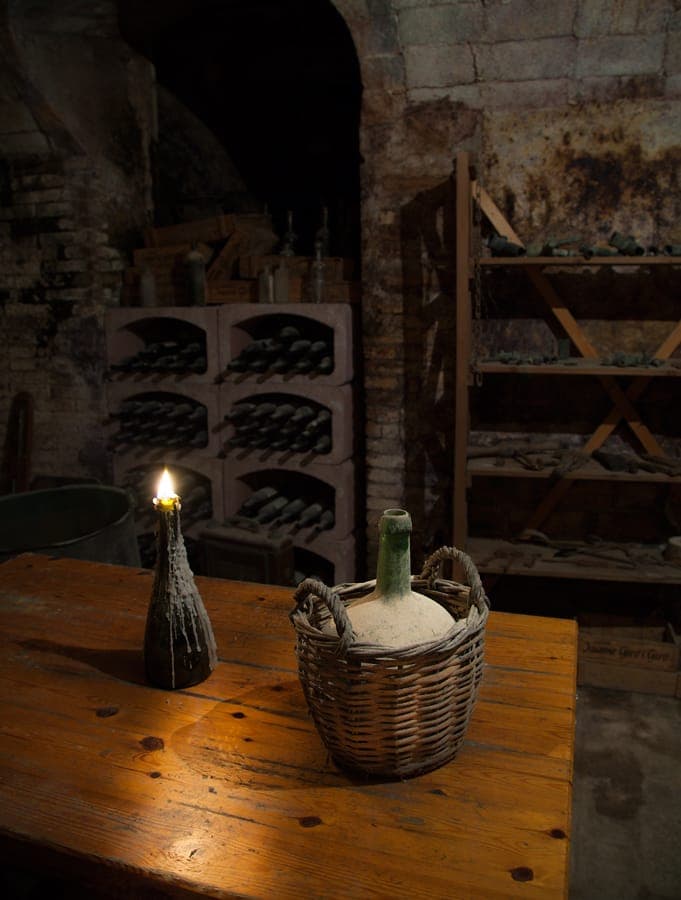 Candle in a wax-covered bottle and wicker jug on a table in a cellar.
